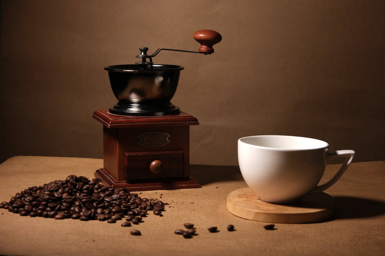 A vintage coffee grinder, coffee beans, and a white mug on a wooden surface, rich with warm tones.